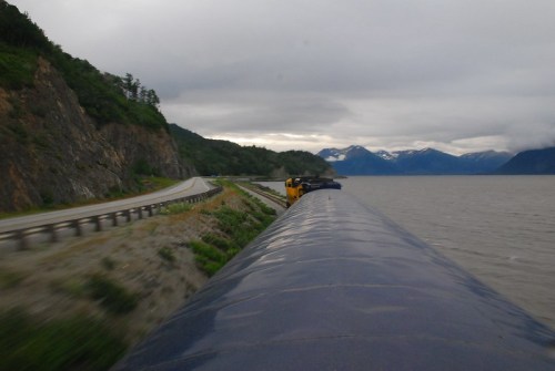 A scenic view of a road alongside a body of water, with mountains in the background under a cloudy sky.