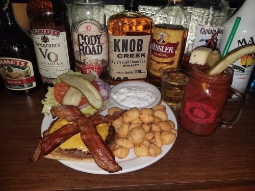 A plate with a cheeseburger topped with bacon, sides of coleslaw and fried cheese balls, and a cocktail in a jar.