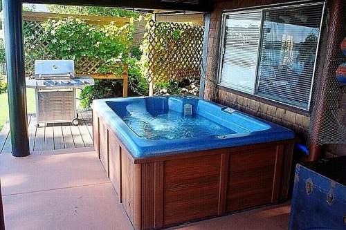 A blue hot tub on a patio with a grill and greenery in the background.