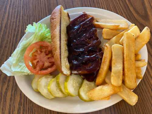 A plate with a BBQ pork sandwich, lettuce, tomato, pickles, and golden fries on a wooden table.