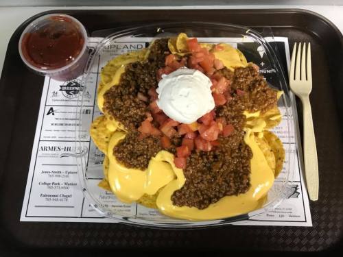 A tray of nachos topped with cheese, chili, diced tomatoes, and a dollop of sour cream, served with salsa on the side.