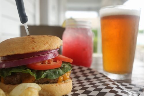 A close-up of a burger with lettuce, tomato, and onion, alongside a drink and a glass of beer on a table.