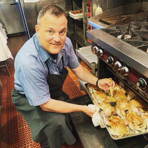 A chef pulls a tray of roasted chicken from an oven in a busy kitchen, smiling while holding a brush.