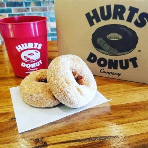 Two sugar-coated donuts on a napkin, with a red cup and a Hurts Donut Company box in the background.