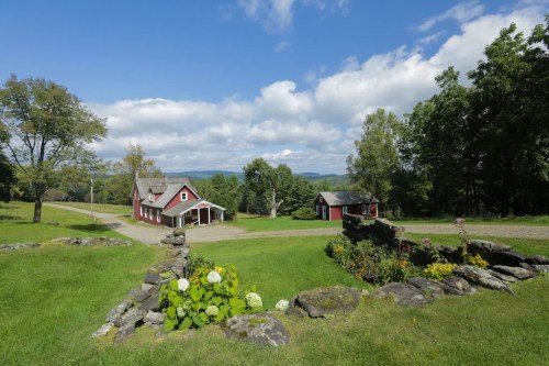 A scenic view of a red farmhouse and a small shed, surrounded by greenery and mountains under a blue sky with clouds.