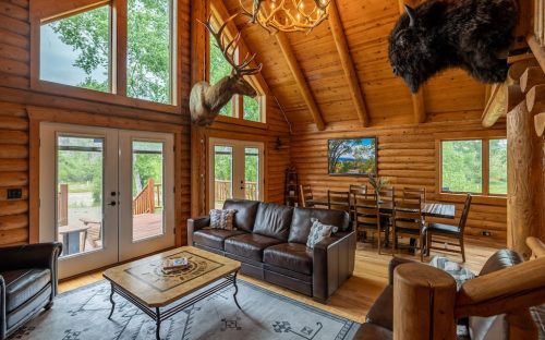 Cozy log cabin interior with a leather sofa, dining area, and mounted animal heads, surrounded by large windows.