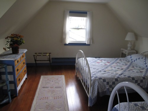 Cozy attic bedroom with two beds, a dresser, and a window, featuring a quilt and warm wooden floors.