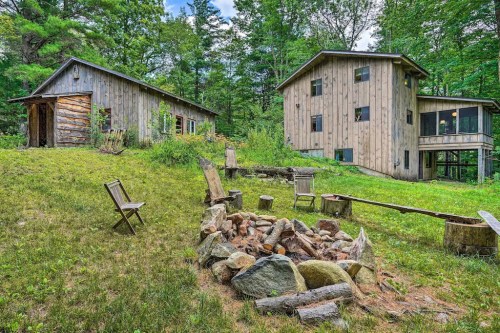 A rustic outdoor scene featuring two wooden buildings and a stone fire pit surrounded by greenery.