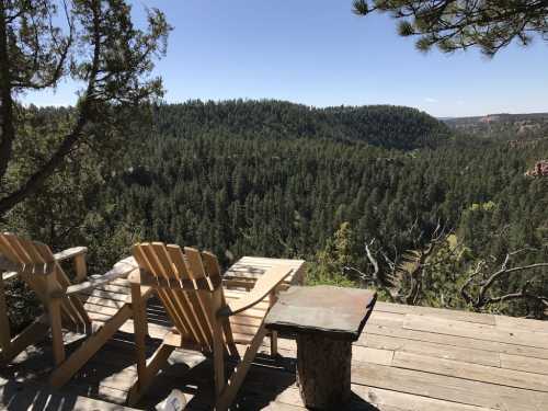 Two wooden chairs on a deck overlooking a lush green forest and distant hills under a clear blue sky.