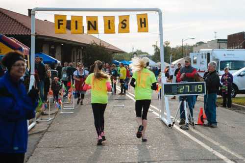 Two runners in bright shirts approach the finish line, with a timer showing 27.19 seconds and a crowd cheering.