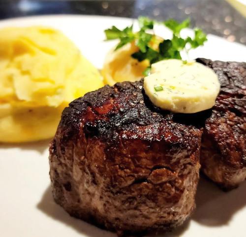 A close-up of two grilled steak medallions topped with herb butter, served with creamy mashed potatoes and garnished with parsley.