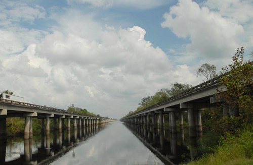 A wide canal flanked by two elevated highways under a cloudy sky, reflecting the scenery in the water below.