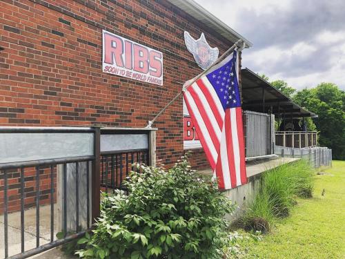 A brick building with a "RIBS" sign and an American flag hanging outside, surrounded by greenery.