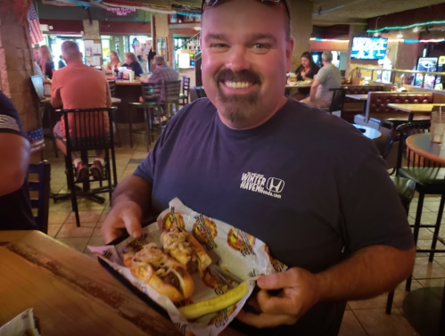 A smiling man holds a plate of hot dogs topped with onions, sitting in a lively restaurant.