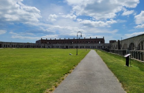 A wide pathway leads to a historic fort surrounded by green grass under a partly cloudy sky.