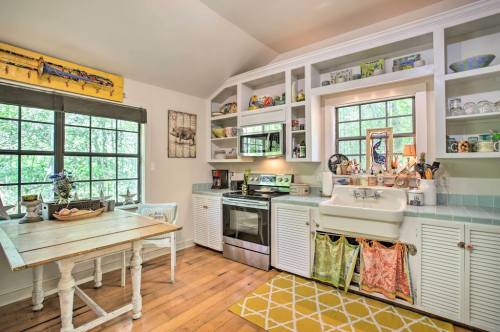 Bright kitchen with a farmhouse sink, stainless steel appliances, open shelves, and a wooden table, surrounded by greenery.