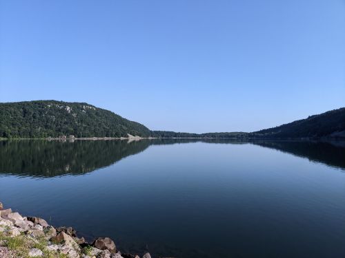 A calm lake surrounded by green hills under a clear blue sky, reflecting the landscape on its surface.
