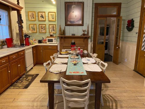 A cozy kitchen with a large wooden table set for a meal, surrounded by cabinets and decorative wall art.