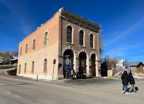 Historic brick building with arched windows, a bell outside, and two people walking on the street under a blue sky.