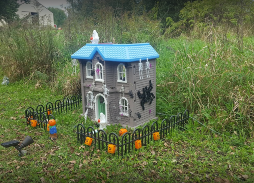 A spooky toy house with skeletons and pumpkins, surrounded by a black fence in a grassy area.