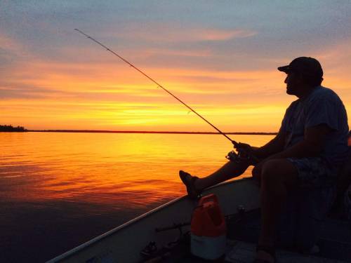 A person fishing from a boat at sunset, with vibrant orange and purple skies reflecting on the calm water.