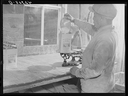 A person pours a substance from a container into a box on a scale in a rustic setting.