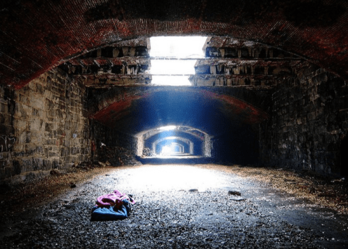 An abandoned, dimly lit tunnel with stone walls and a colorful blanket on the ground, illuminated by light from above.