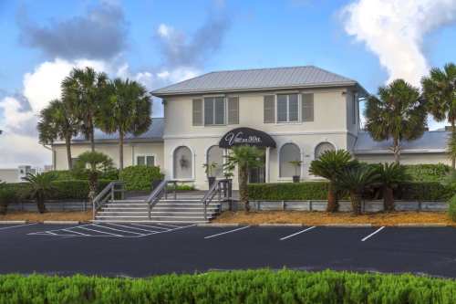 A modern two-story building with a metal roof, surrounded by palm trees and greenery, featuring a welcoming entrance.