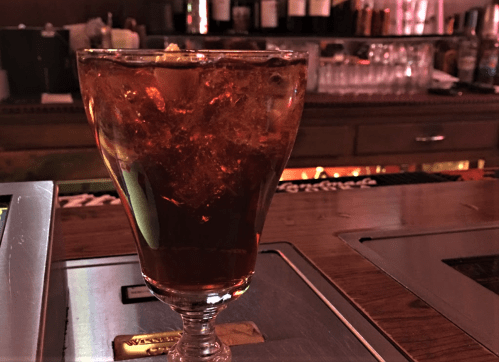 A glass of dark soda with ice and a slice of lemon, sitting on a bar counter with blurred bottles in the background.