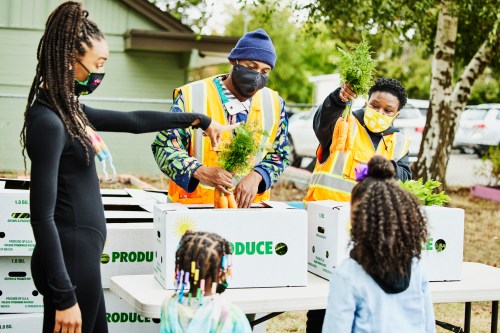 A group of people in masks distribute fresh produce to children at an outdoor community event.