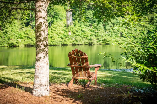 A wooden Adirondack chair sits by a serene lake, surrounded by trees and greenery, with a gentle breeze moving the wind chimes.