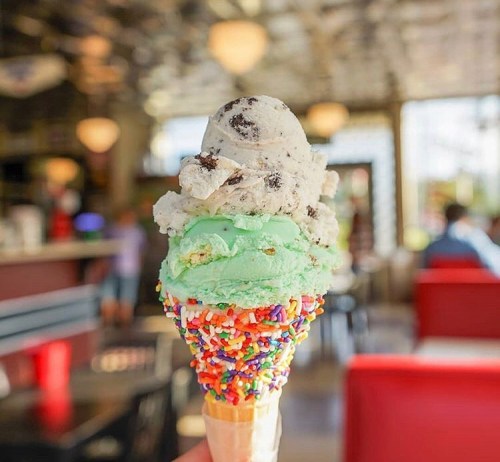 A colorful ice cream cone with mint, cookies and cream, and sprinkles, set against a vibrant ice cream shop background.