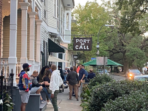 A lively street scene with people gathered outside a restaurant called "Port of Call" under a tree-lined street.