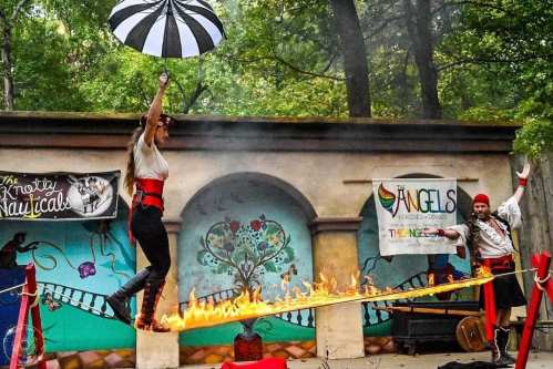 A performer balances on a tightrope over flames, holding an umbrella, while a pirate watches in a colorful outdoor setting.