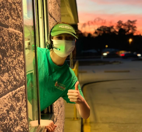 A smiling employee in a green shirt and mask gives a thumbs up from a drive-thru window at sunset.
