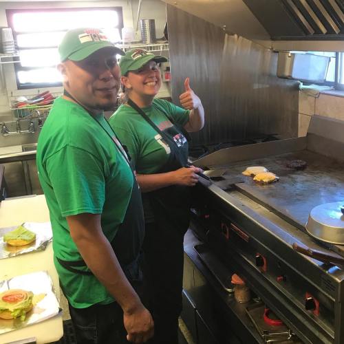 Two chefs in green shirts smile at the camera while cooking on a grill in a kitchen. One gives a thumbs up.