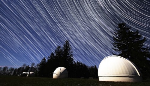Star trails swirl above observatories surrounded by trees in a night sky.