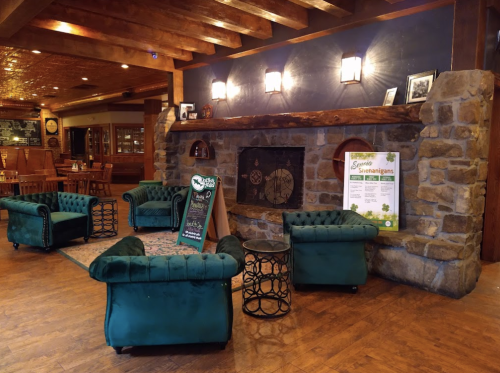 Cozy lounge area with green tufted chairs, a stone fireplace, and wooden beams, featuring a menu board and warm lighting.