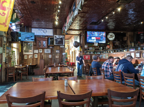 A cozy restaurant interior with wooden tables, wall decor, and patrons enjoying their meals. TVs are mounted on the walls.