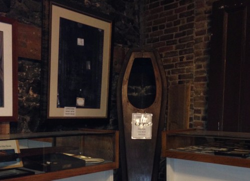 A wooden coffin displayed in a museum, surrounded by framed pictures and glass cases.
