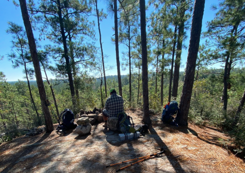 A hiker sits on a rocky outcrop surrounded by trees, with backpacks and hiking poles nearby, overlooking a scenic view.