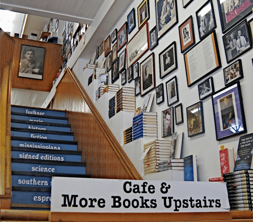 Staircase leading to an upstairs cafe, lined with framed photos and bookshelves, with a sign for "Cafe & More Books Upstairs."