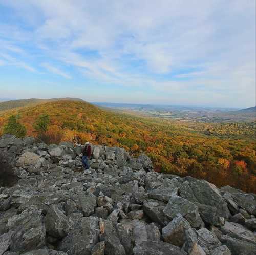 A person walks among large rocks on a hillside, overlooking a colorful valley and distant mountains under a blue sky.