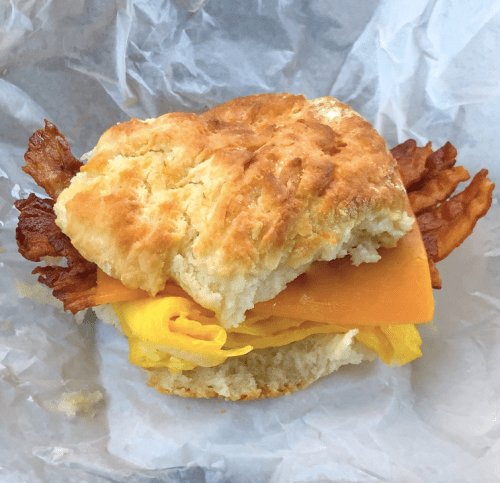 A biscuit sandwich filled with crispy bacon, cheddar cheese, and scrambled eggs, wrapped in parchment paper.