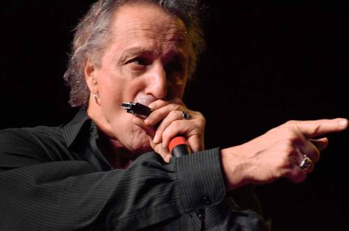 A musician passionately playing harmonica while singing on stage, gesturing with one hand.