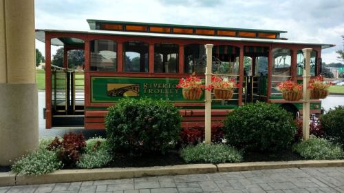A green and wooden trolley with "Riverton Trolley" signage parked near flower planters and shrubs.