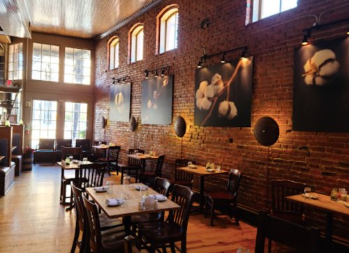 Cozy restaurant interior with wooden tables, brick walls, and large cotton flower artworks. Natural light from windows.