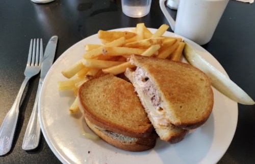 A plate with a grilled sandwich, golden fries, and a slice of pickle, set on a black table.