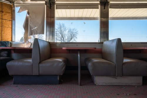 Abandoned diner interior with empty booths and a broken window, showcasing peeling walls and a dusty floor.