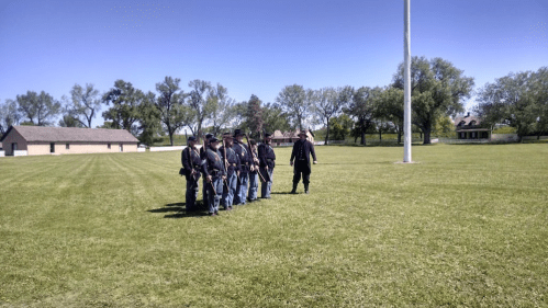A group of soldiers in historical uniforms marching across a grassy field with buildings in the background.